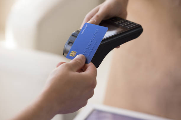 istockphoto-1473718519-612×612 Hand of bank cardholder paying bill in cafe, applying blue credit card with chip at wireless payment terminal held by waitress, using electronic transaction banking technology. Clopped shot, close up
